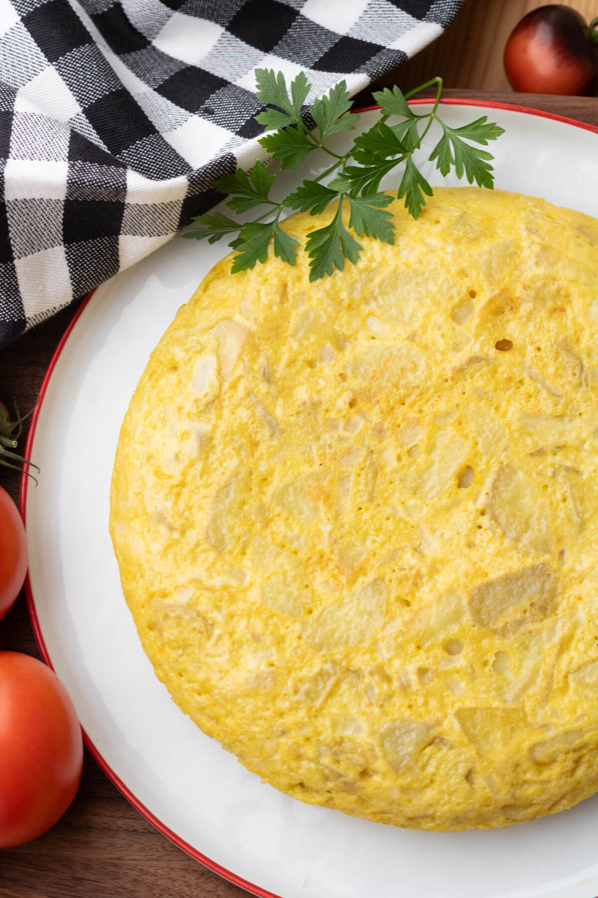 A perfect round yellow Spanish Tortilla or Tortilla de Patatas on a white plate with a red rim surrounded by tomatoes.