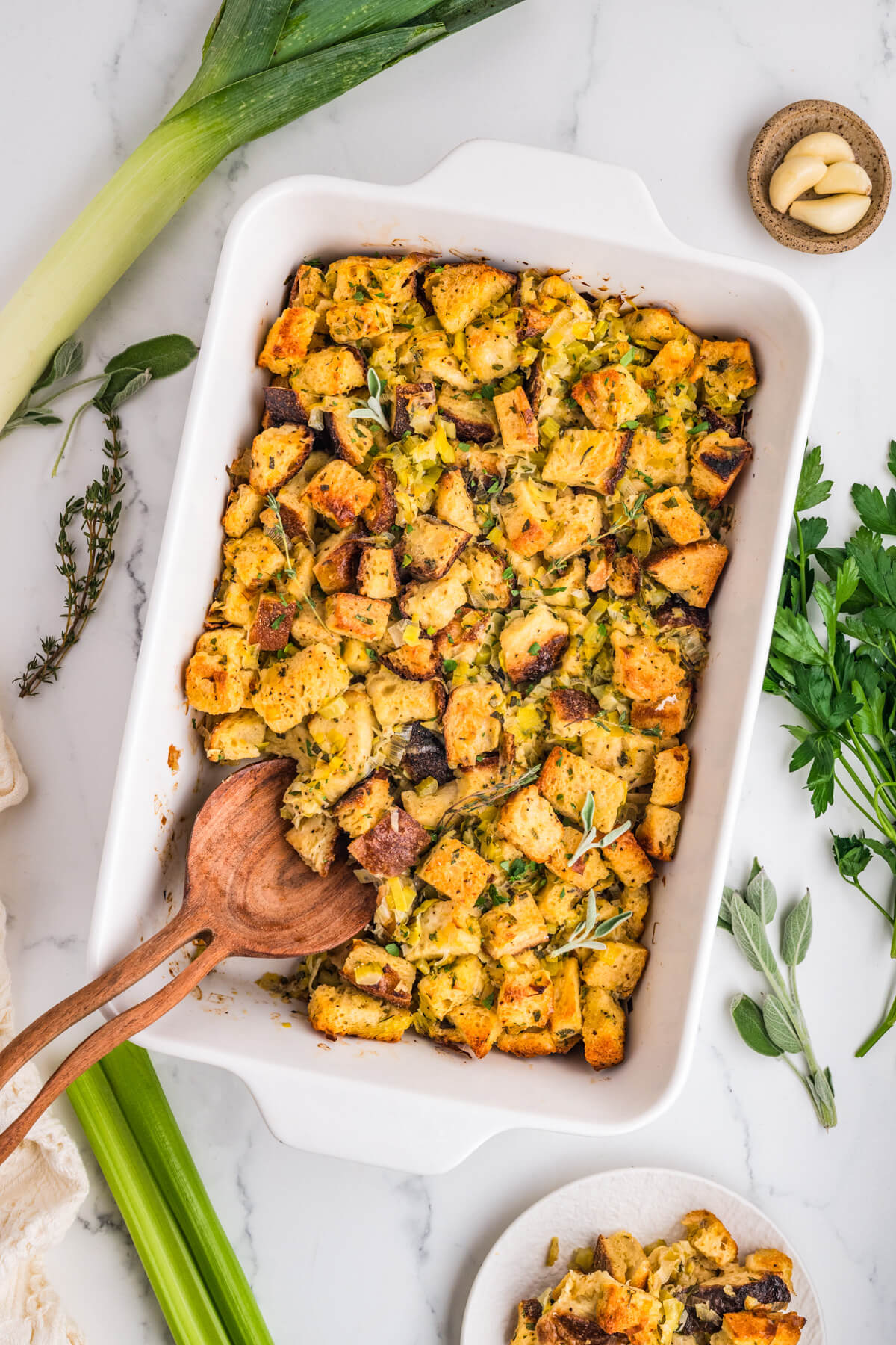 A white baking dish filled with golden baked sourdough bread cubes seasoned with fresh herbs and aromatics.