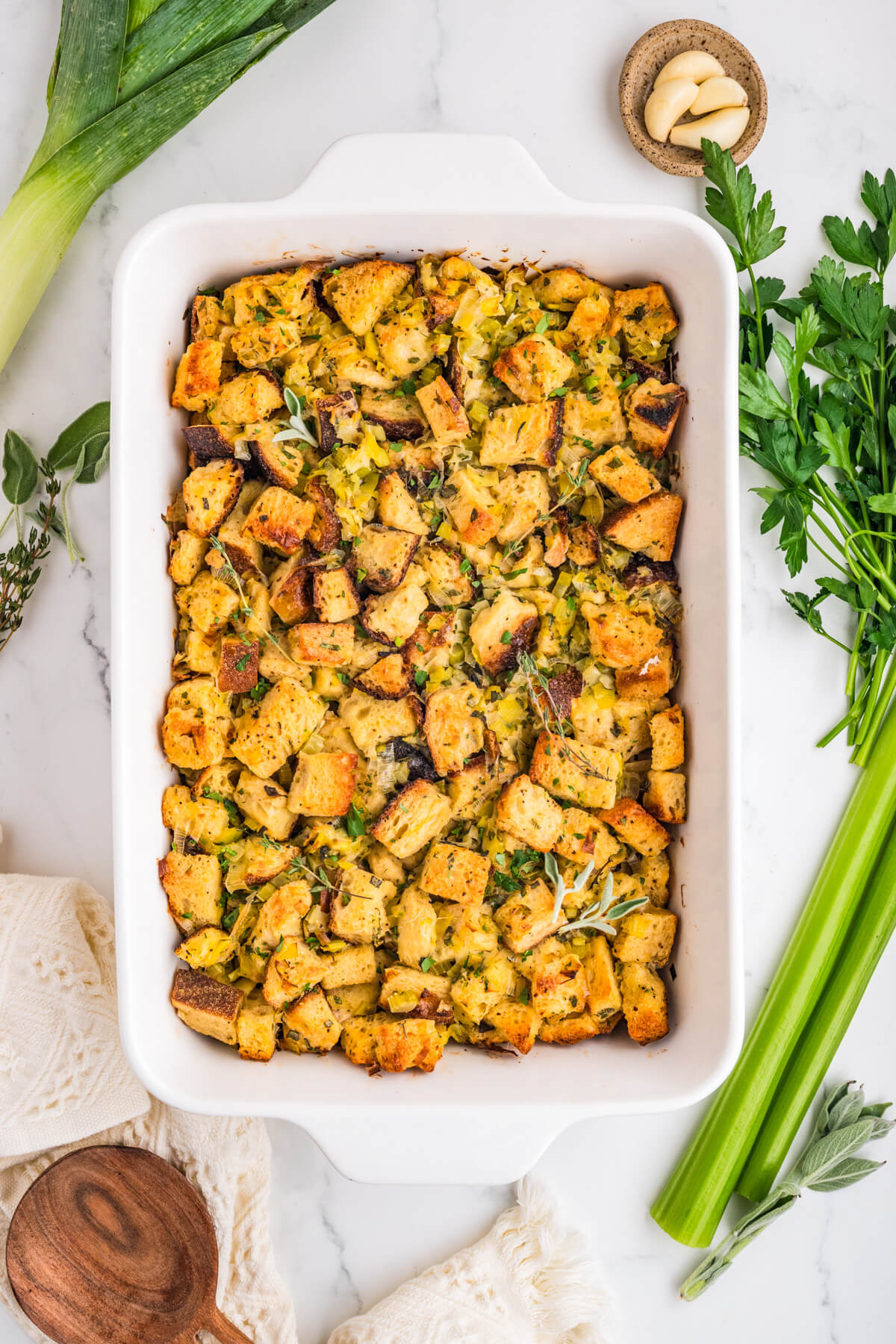 A white baking dish filled with golden baked sourdough bread cubes seasoned with fresh herbs and aromatics.