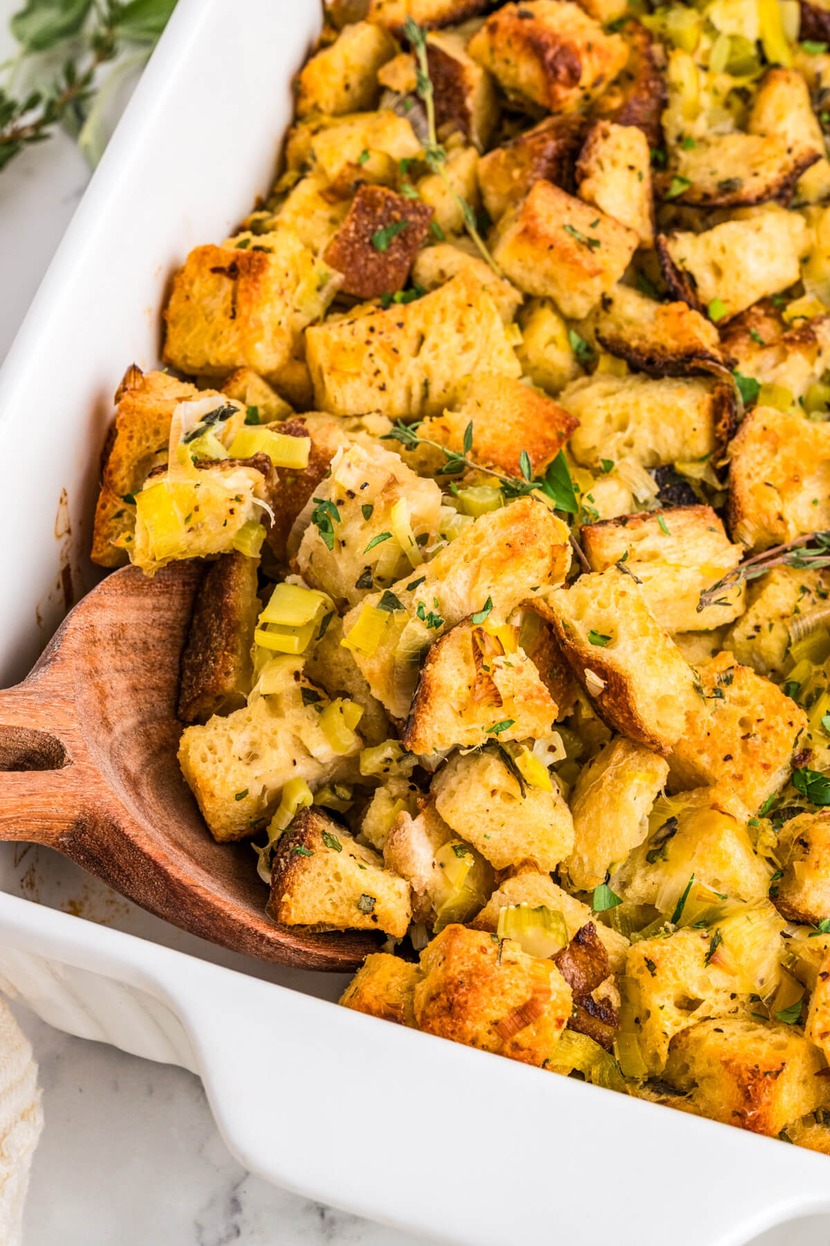 A white baking dish filled with golden baked sourdough bread cubes seasoned with fresh herbs and aromatics.