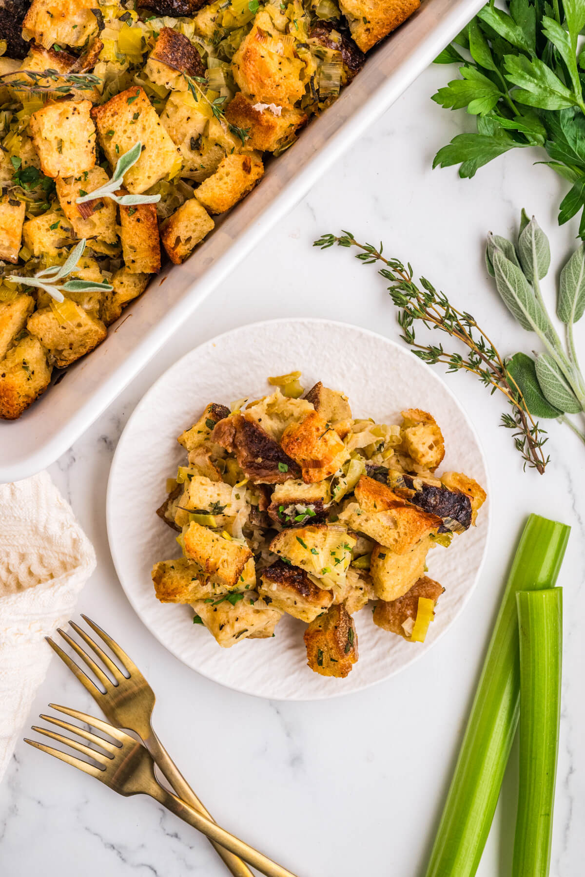 A white plate containing golden baked sourdough bread cubes seasoned with fresh herbs and aromatics on a dinner table.