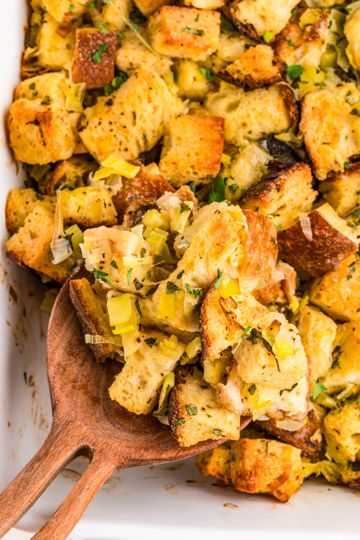 A white baking dish full of golden baked sourdough stuffing with green herbs and aromatics.
